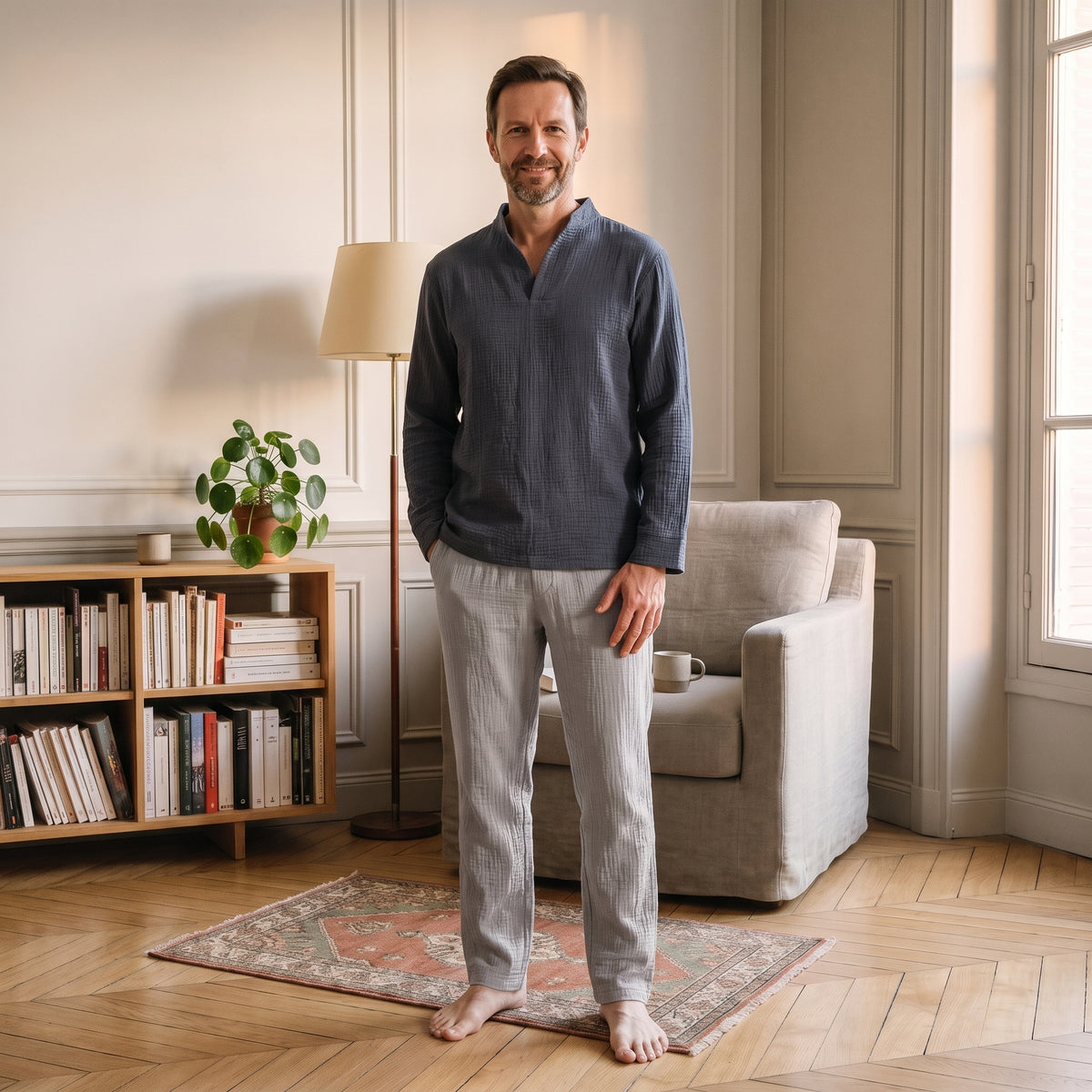 Homme en pyjama de double gaze de coton, haut bleu ardoise et pantalon gris clair, debout dans le coin lecture