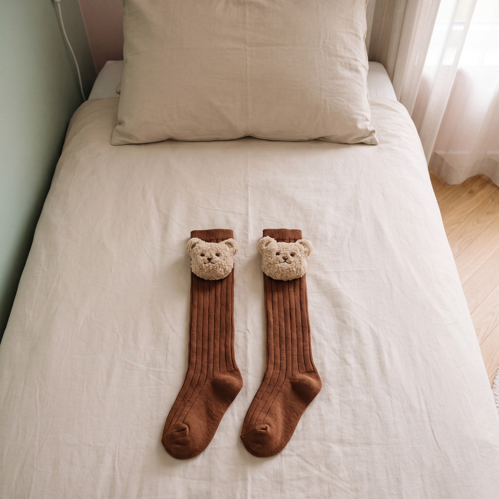Chaussettes montantes camel pour bébé avec un ourson en peluche, posées sur un lit d'enfant