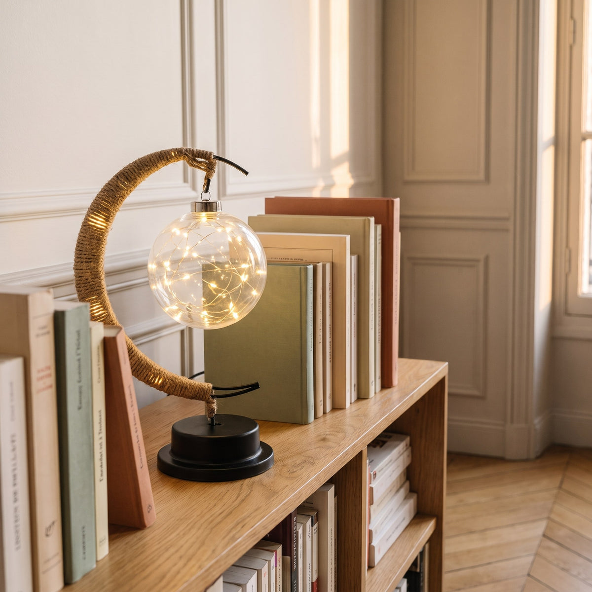 La lampe posée sur une étagère de bibliothèque, socle noir à plat entre les livres.