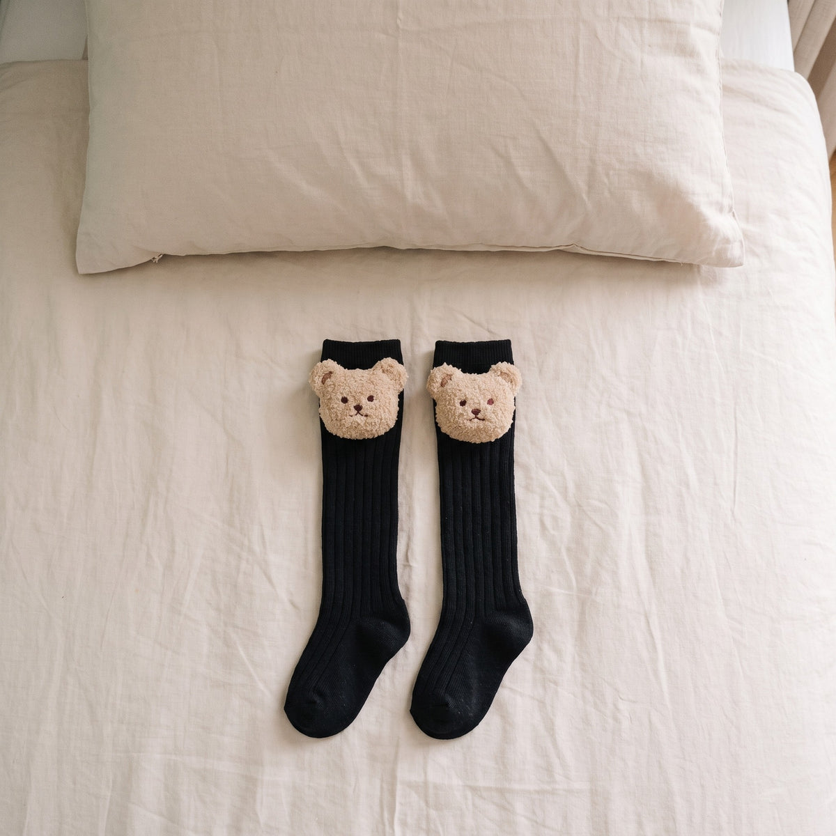 Chaussettes montantes noires pour bébé avec un ourson en peluche, posées sur un lit d'enfant