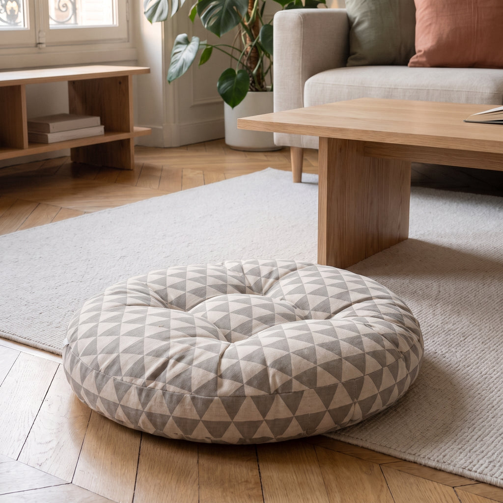 Coussin de sol rond à triangles gris sur fond écru, posé au sol près d'une table basse