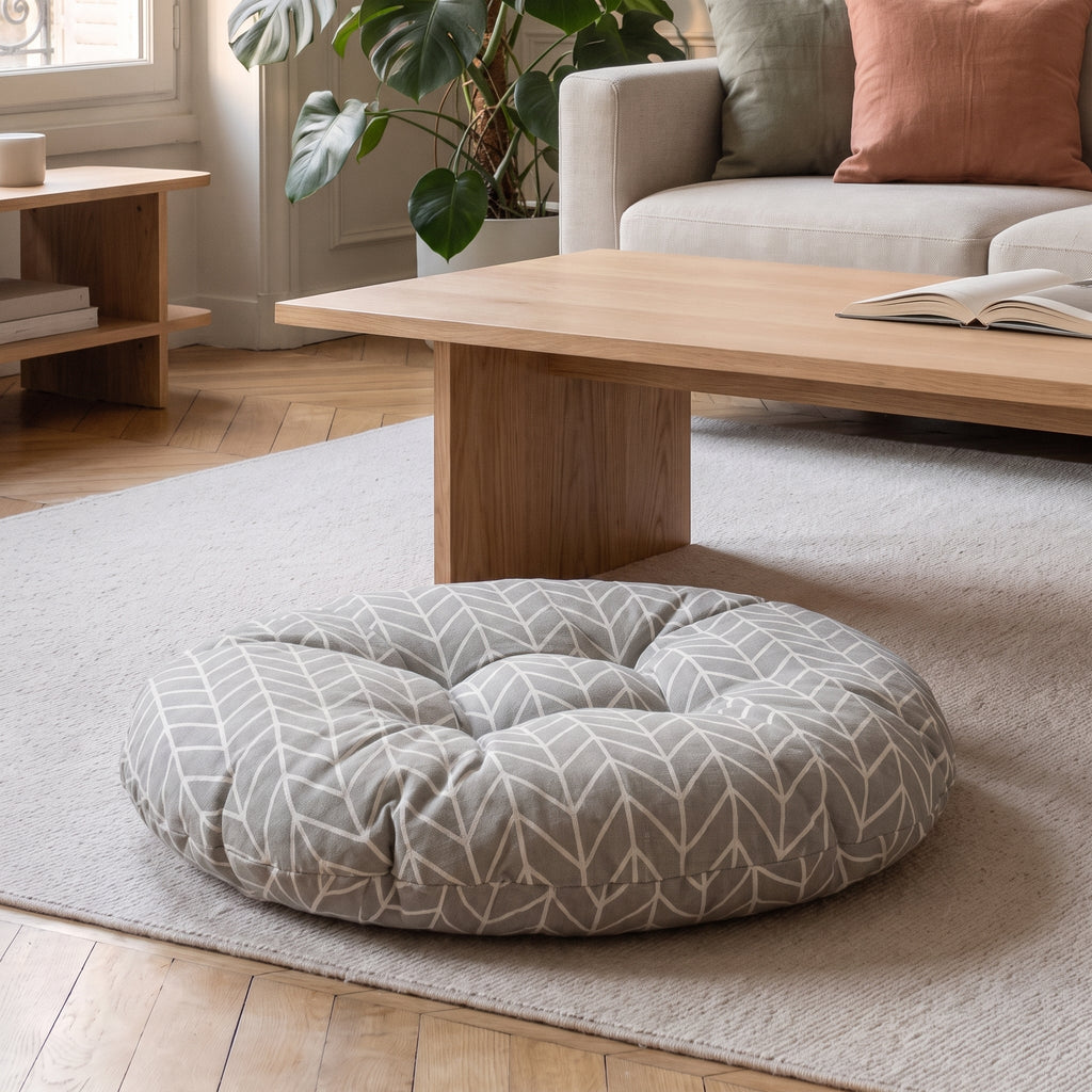 Coussin de sol rond à chevrons gris clair, posé au sol devant une table basse en bois