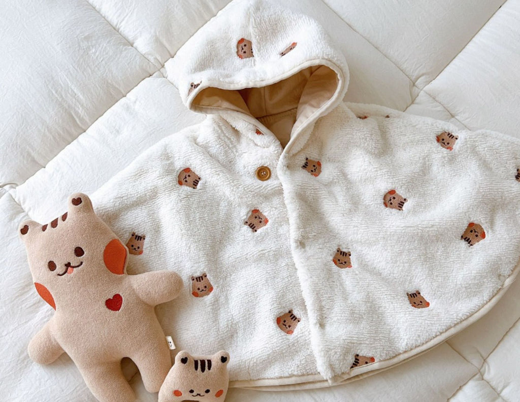 La cape motif tigre brun posee a plat sur un lit, capuche ouverte sur sa doublure beige, une peluche posee a cote