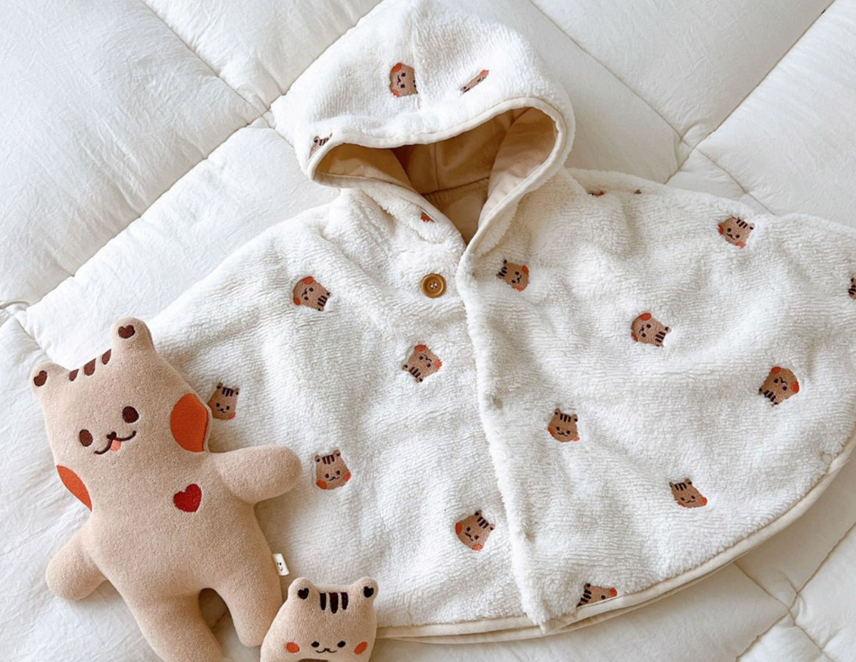 La cape motif tigre brun posee a plat sur un lit, capuche ouverte sur sa doublure beige, une peluche posee a cote