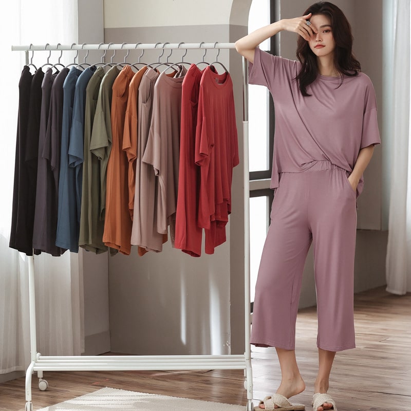 Femme debout accoudée à un portant, porant un ensemble fluide, cocooning, rose et des sandales. Cette tenue dans différentes couleurs est suspendue sur le portant.