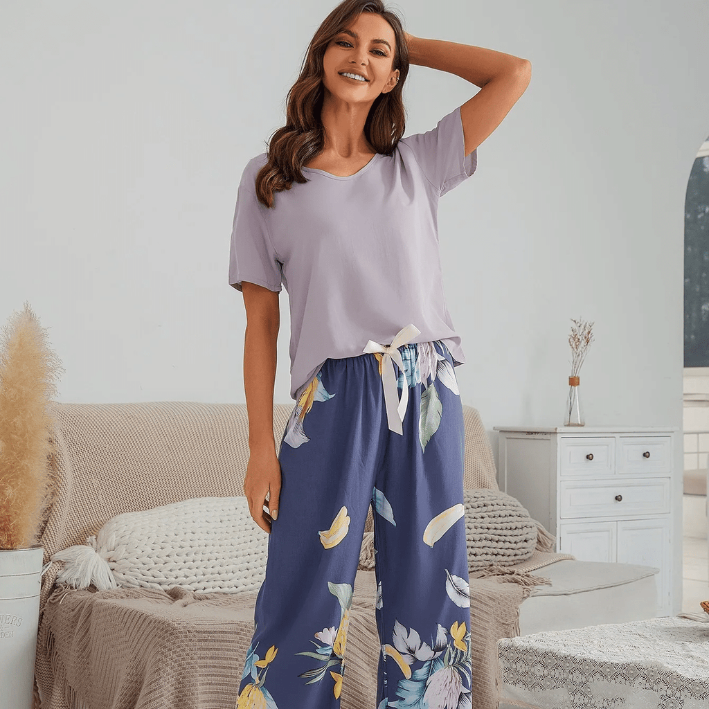 Pyjama femme, haut lilas et pantalon large feuillage bleu, debout dans une chambre claire, le bras derrière la tête.