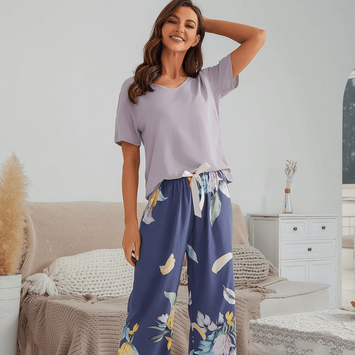 Pyjama femme, haut lilas et pantalon large feuillage bleu, debout dans une chambre claire, le bras derrière la tête.