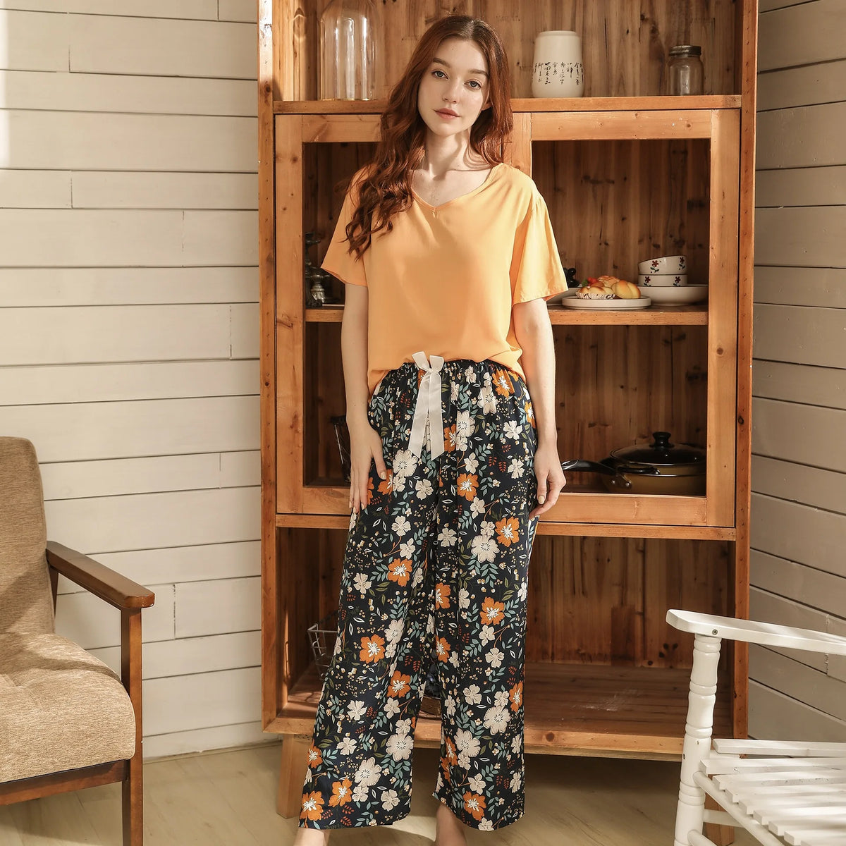 Pyjama femme, haut abricot et pantalon large fleurs blanches marine, debout devant un vaisselier en bois, de face.