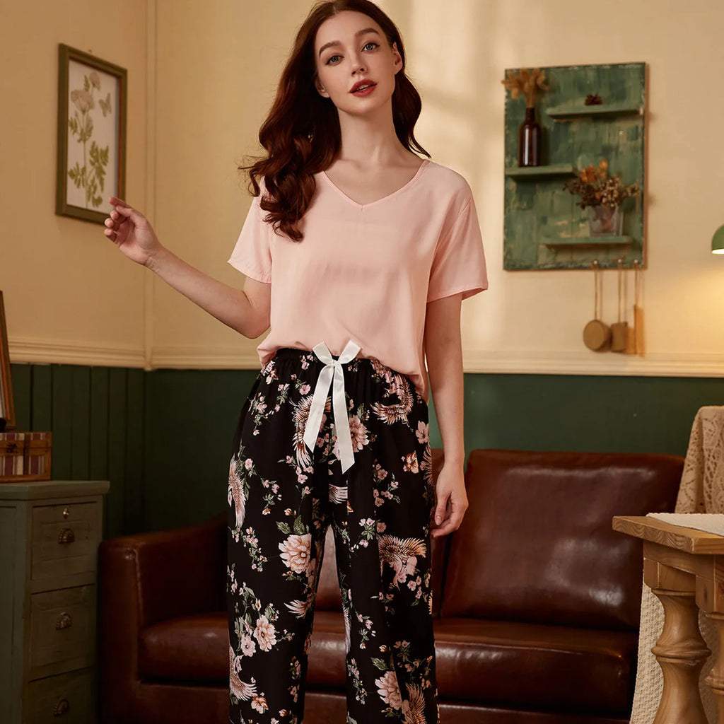 Pyjama femme, haut rose et pantalon large petites fleurs noir, debout dans un salon vert, un canapé de cuir derrière elle.