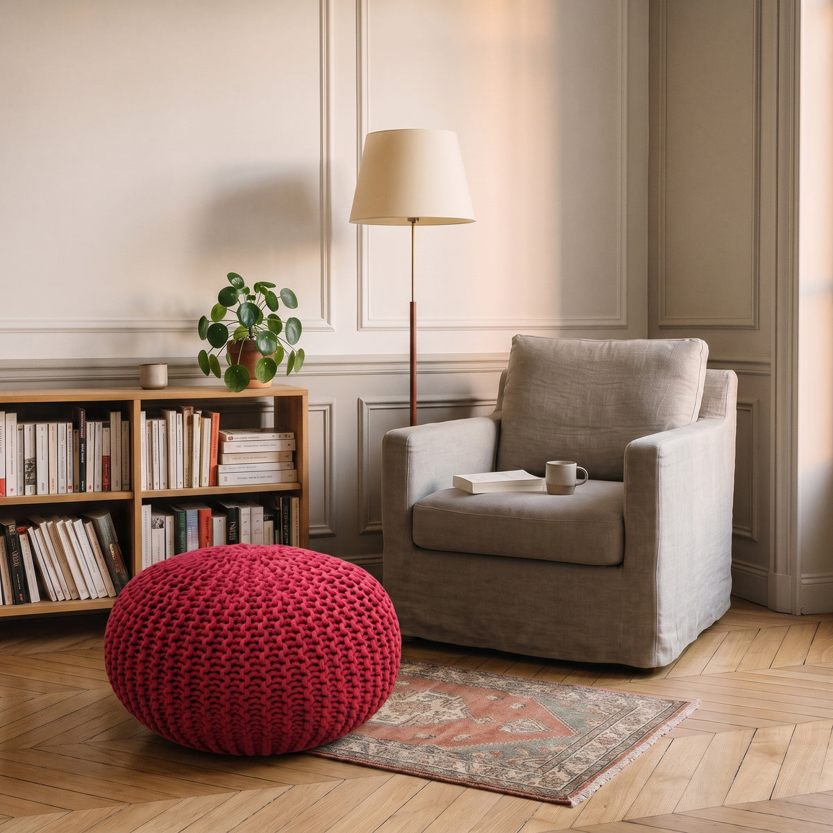 Pouf rond en grosse maille rouge posé sur le parquet devant un fauteuil de lecture en lin clair, à côté d'une bibliothèque basse