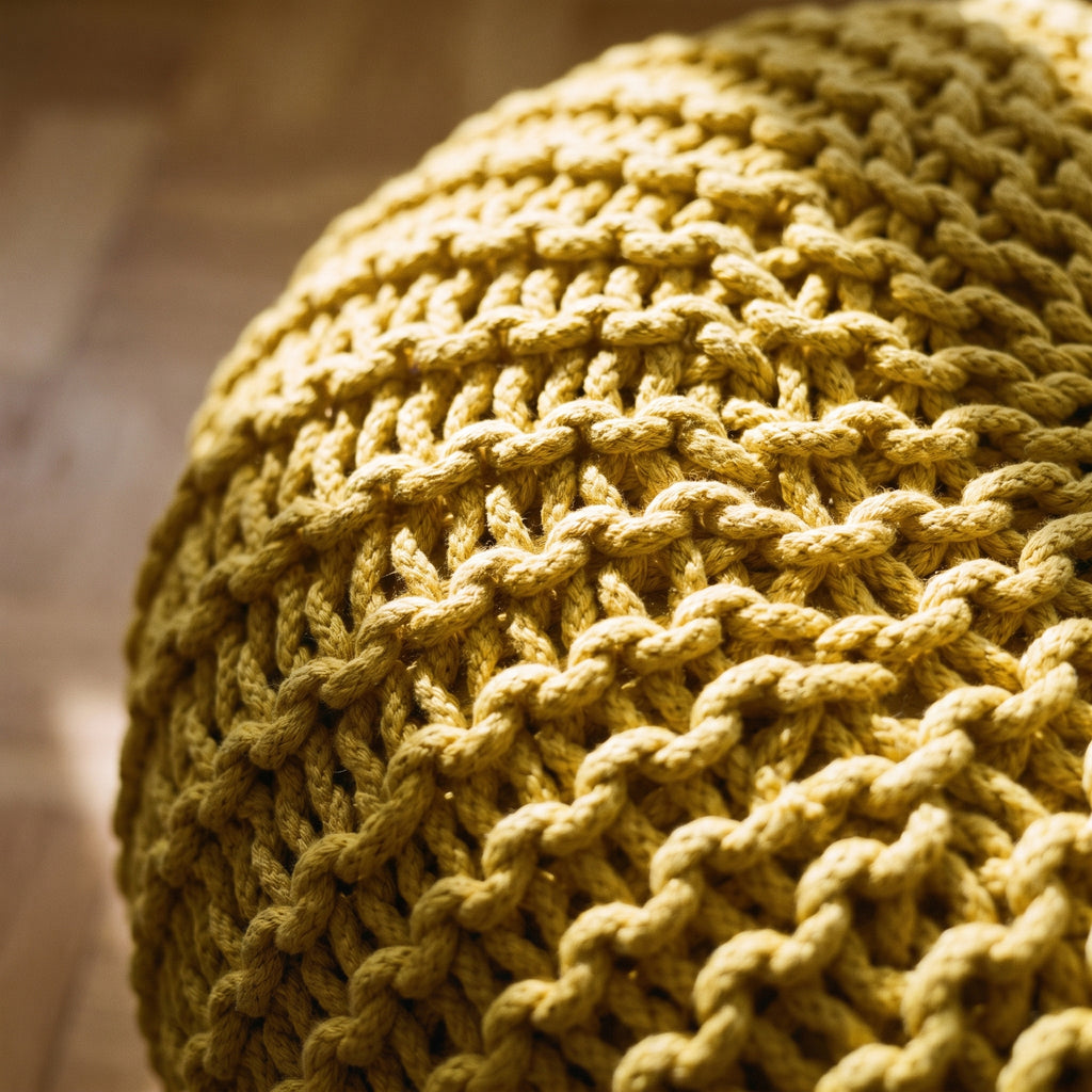 Gros plan sur la grosse maille jaune moutarde du pouf, la lumière rasante allume la crête de chaque rang et creuse l'ombre entre deux rangs