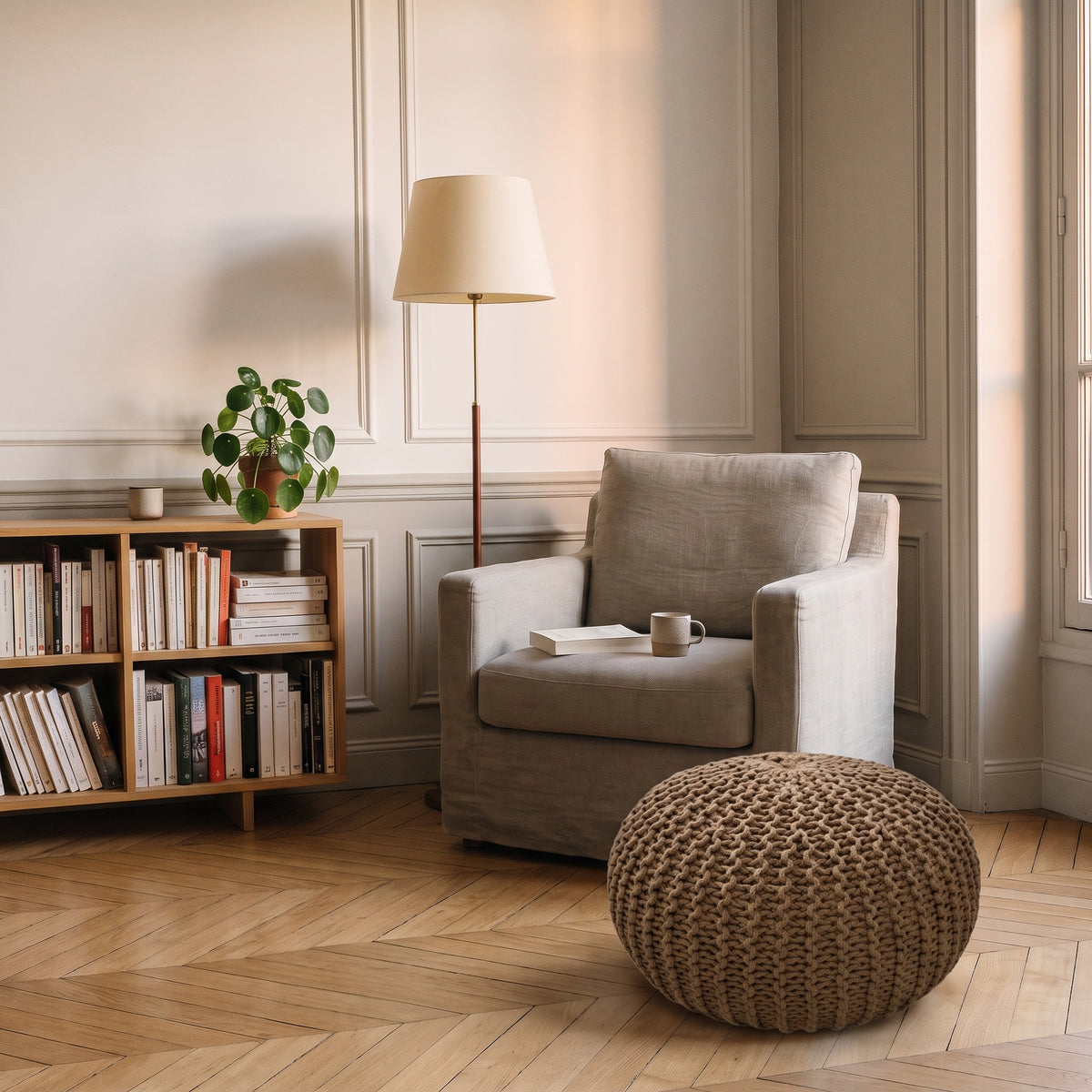 Pouf rond en grosse maille marron posé sur le parquet en point de Hongrie d'un coin lecture, devant un fauteuil en lin clair