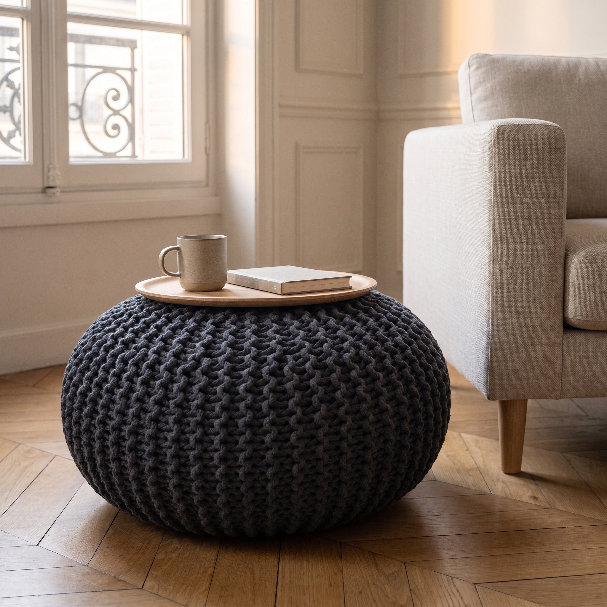 Pouf rond en grosse maille noire utilisé en table d'appoint, un plateau ovale en bois clair posé sur son dessus avec une tasse en grès et un livre Image