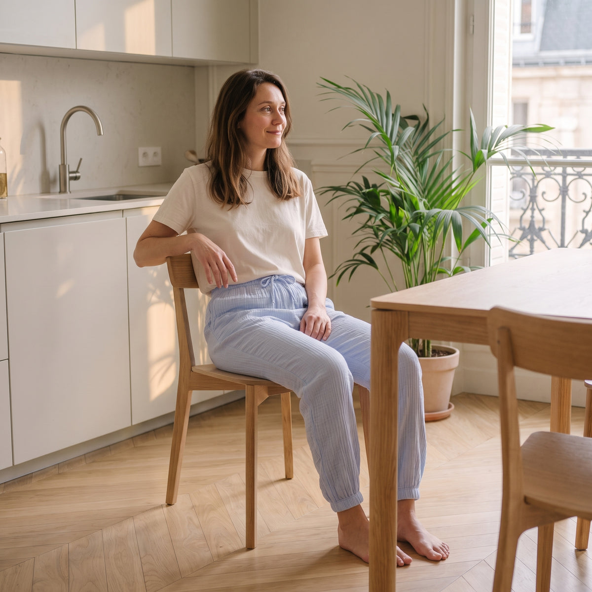 Femme assise à la table du coin repas en pantalon de pyjama bleu ciel et tee-shirt écru, pieds nus Image