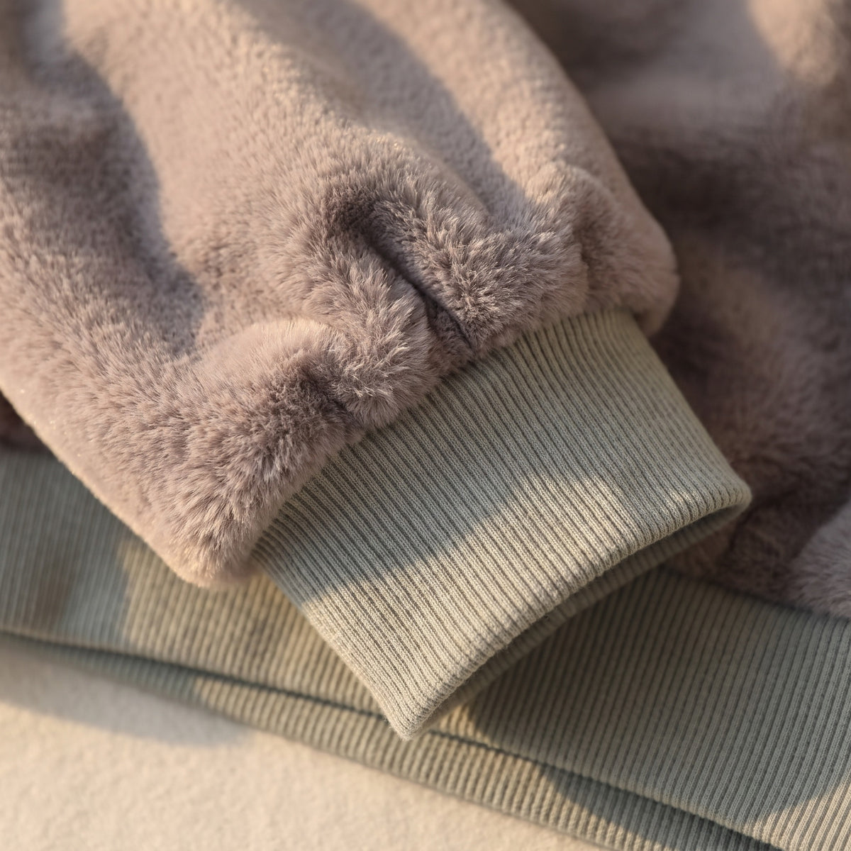 Gros plan sur le bas d une manche grise en lumiere rasante : la peluche fronce sur la bande de maille cotelee vert sauge, dont les cotes se comptent une a une