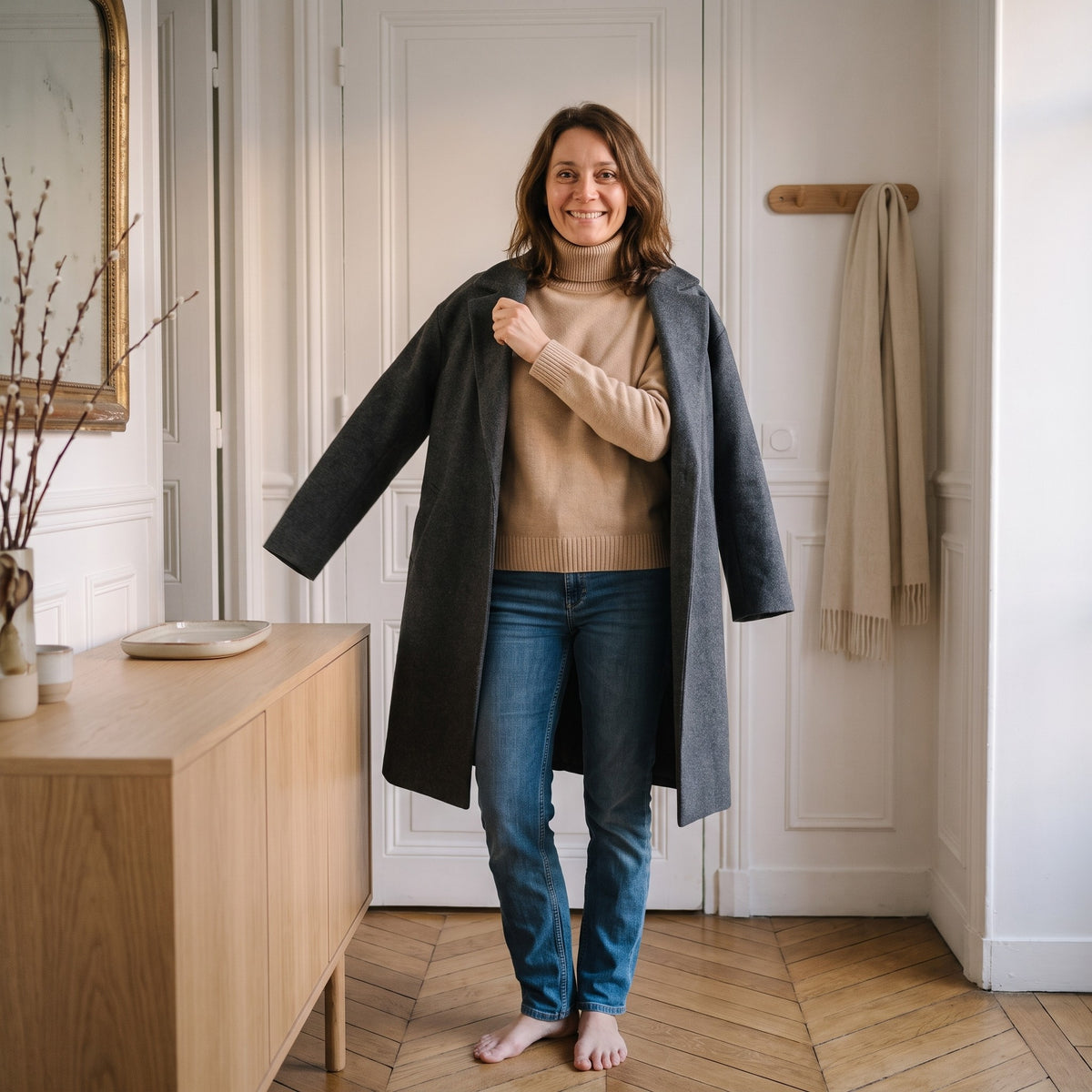 Femme dans son entree qui enfile un manteau de laine gris par-dessus le pull col roule camel, dont le col depasse au-dessus du col du manteau