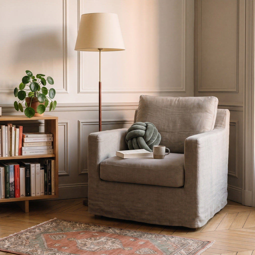 Coussin nœud kaki grisé calé au creux d'un fauteuil de lecture, à côté d'un livre ouvert