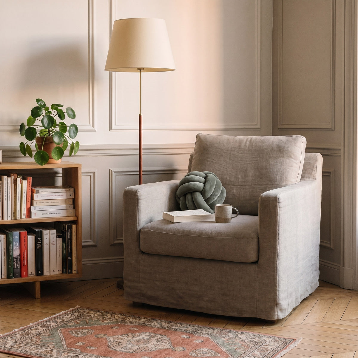 Coussin nœud kaki grisé calé au creux d'un fauteuil de lecture, à côté d'un livre ouvert
