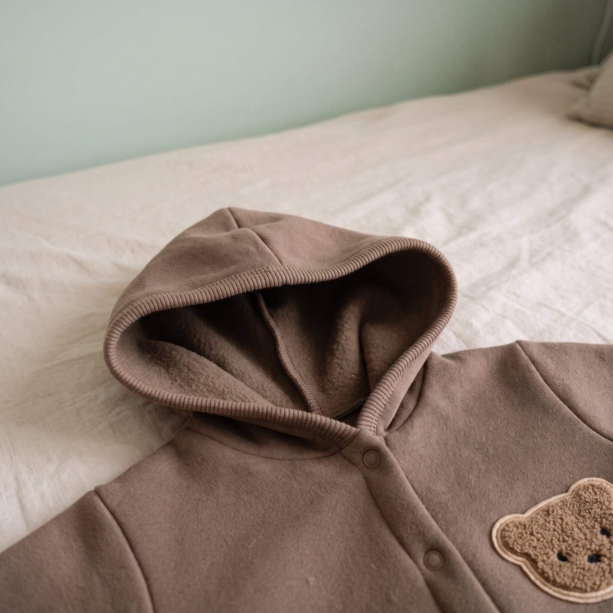 Capuche relevée de la barboteuse bébé marron posée sur un lit d'enfant, doublure polaire visible Image