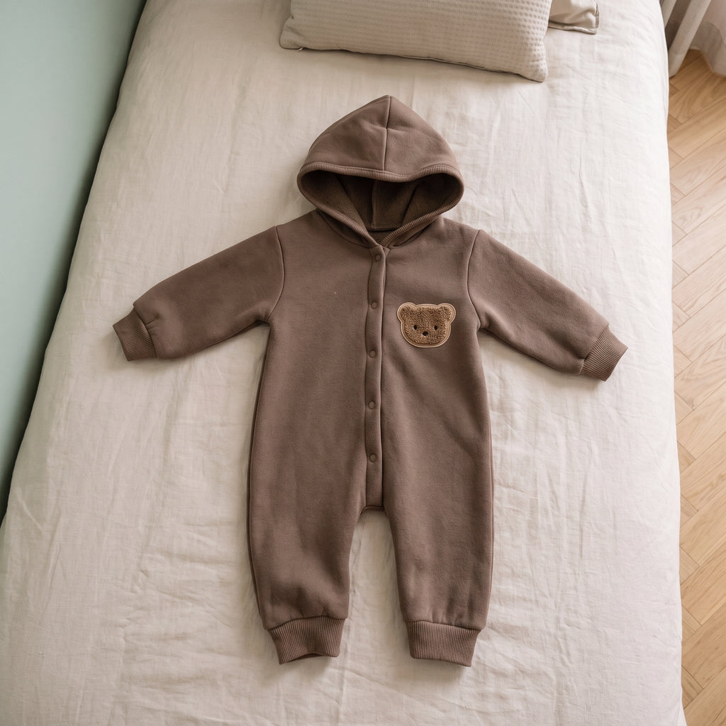 Barboteuse bébé marron à capuche avec écusson ourson, posée à plat sur un lit d'enfant aux draps écrus