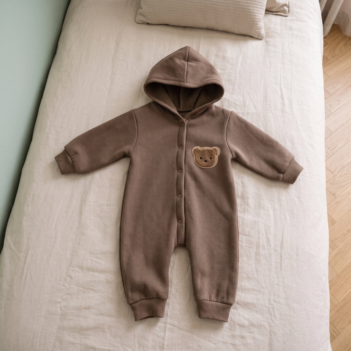 Barboteuse bébé marron à capuche avec écusson ourson, posée à plat sur un lit d'enfant aux draps écrus