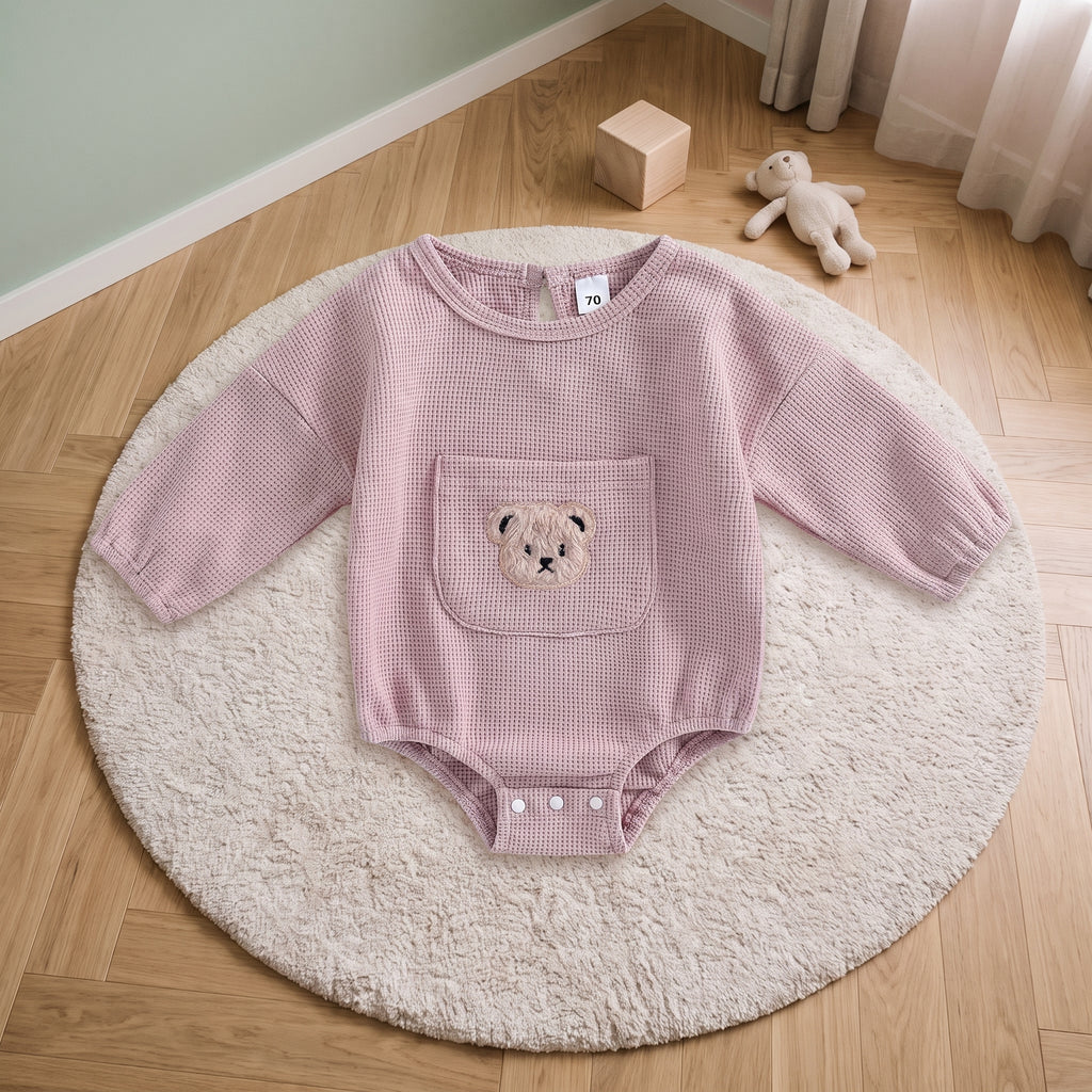 Barboteuse bébé rose poudré en maille gaufrée, posée sur un tapis rond crème