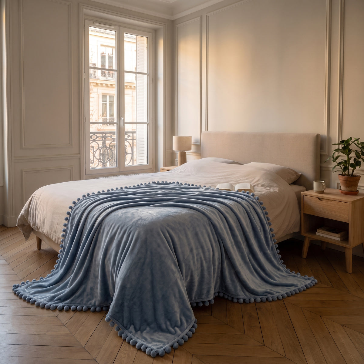 Plaid molletonné bleu gris à pompons déplié sur un lit deux places