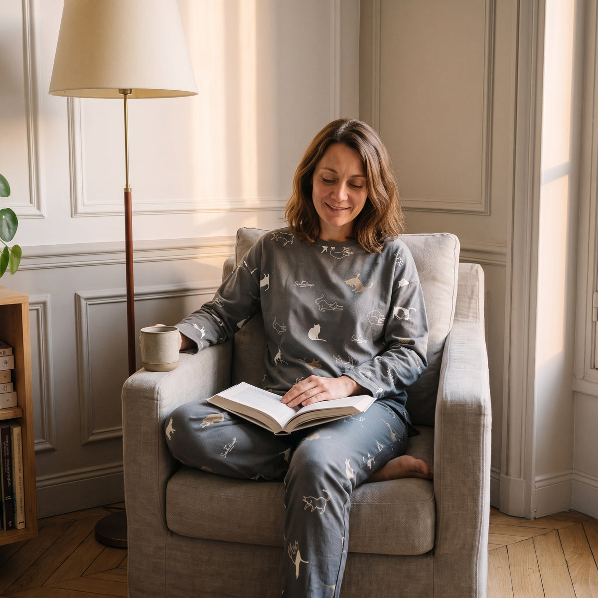 Femme en pyjama gris-vert assise dans le fauteuil de lecture, un livre ouvert sur les genoux et une tasse sur l accoudoir. Image