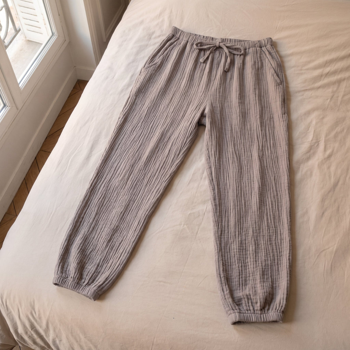 Pantalon de pyjama gris taupe en gaze de coton pose a plat sur le lit, vu du dessus