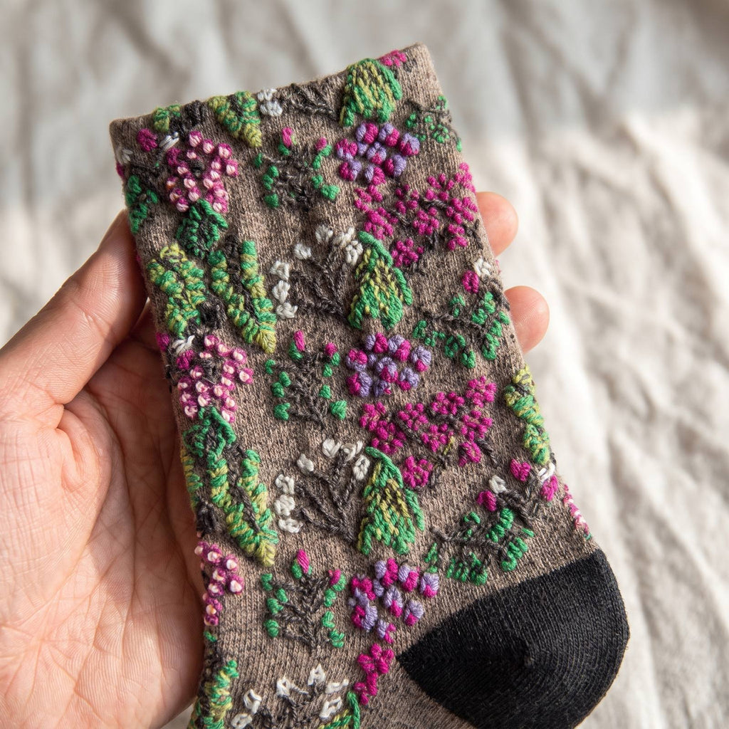 Gros plan sur la tige d'une chaussette fleurie tenue à la main, le semis de corolles et de baies se détache en relief au-dessus de la maille taupe