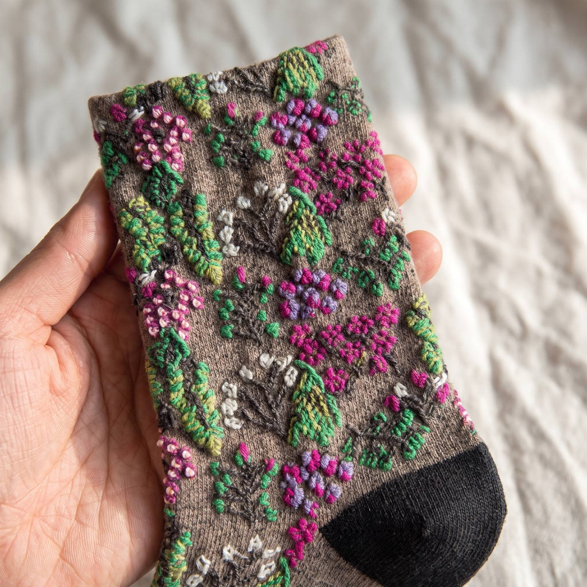 Gros plan sur la tige d'une chaussette fleurie tenue à la main, le semis de corolles et de baies se détache en relief au-dessus de la maille taupe