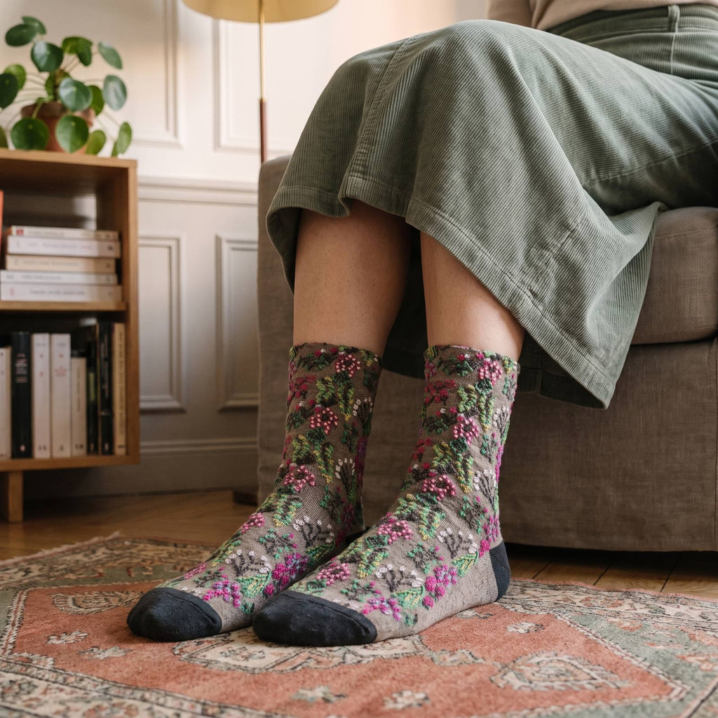 Chaussettes fleuries beige portées, motif en relief vert et rose et pointe anthracite, pieds posés sur un tapis à médaillon dans un coin lecture haussmannien