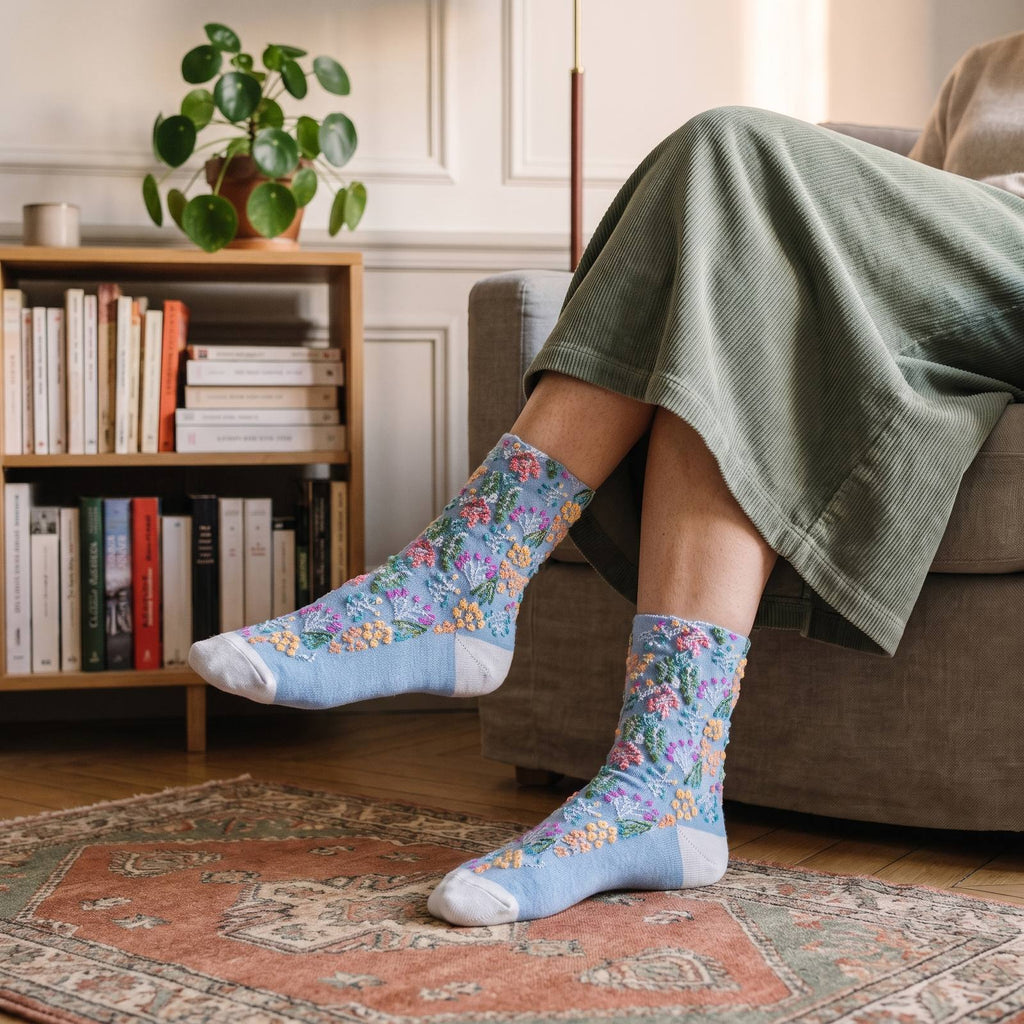 Chaussettes fleuries bleu ciel portées, fleurs abricot et roses et talon blanc, un pied tendu devant une bibliothèque basse en bois clair