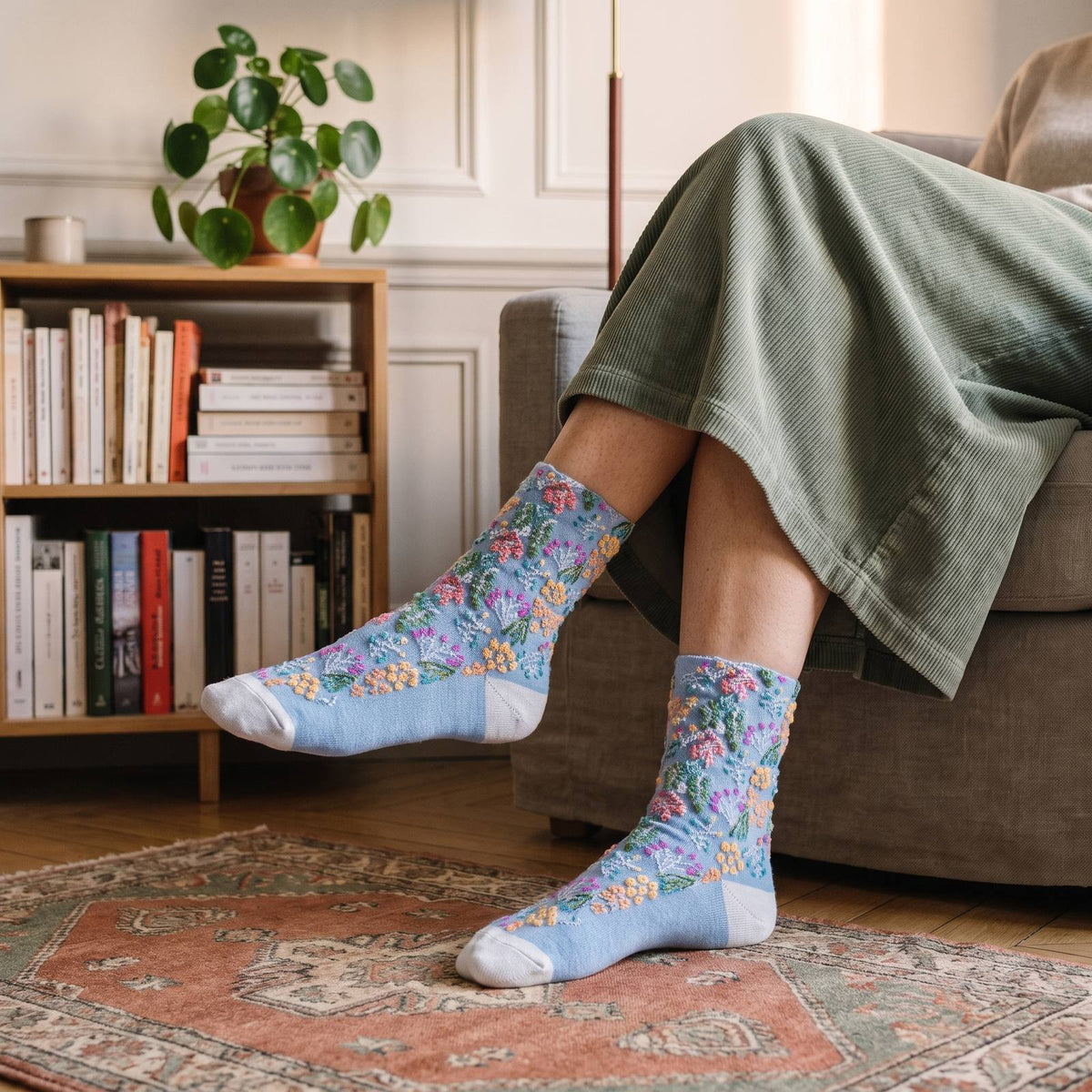 Chaussettes fleuries bleu ciel portées, fleurs abricot et roses et talon blanc, un pied tendu devant une bibliothèque basse en bois clair