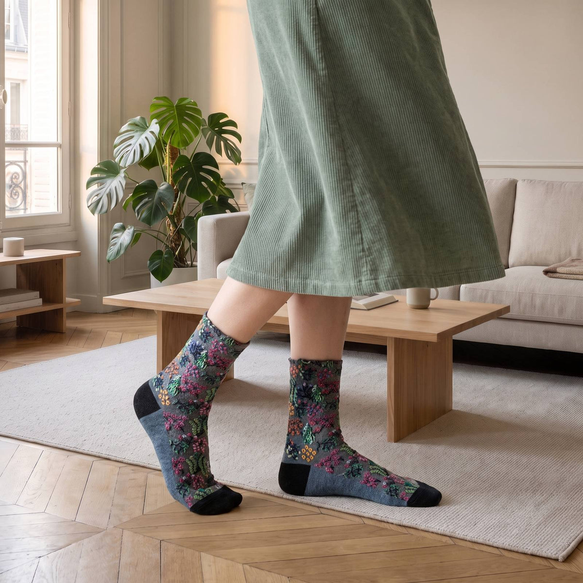 Chaussettes fleuries bleu ardoise portées debout sur un tapis crème, motif orange et vert et pointe noire, salon haussmannien avec fenêtre