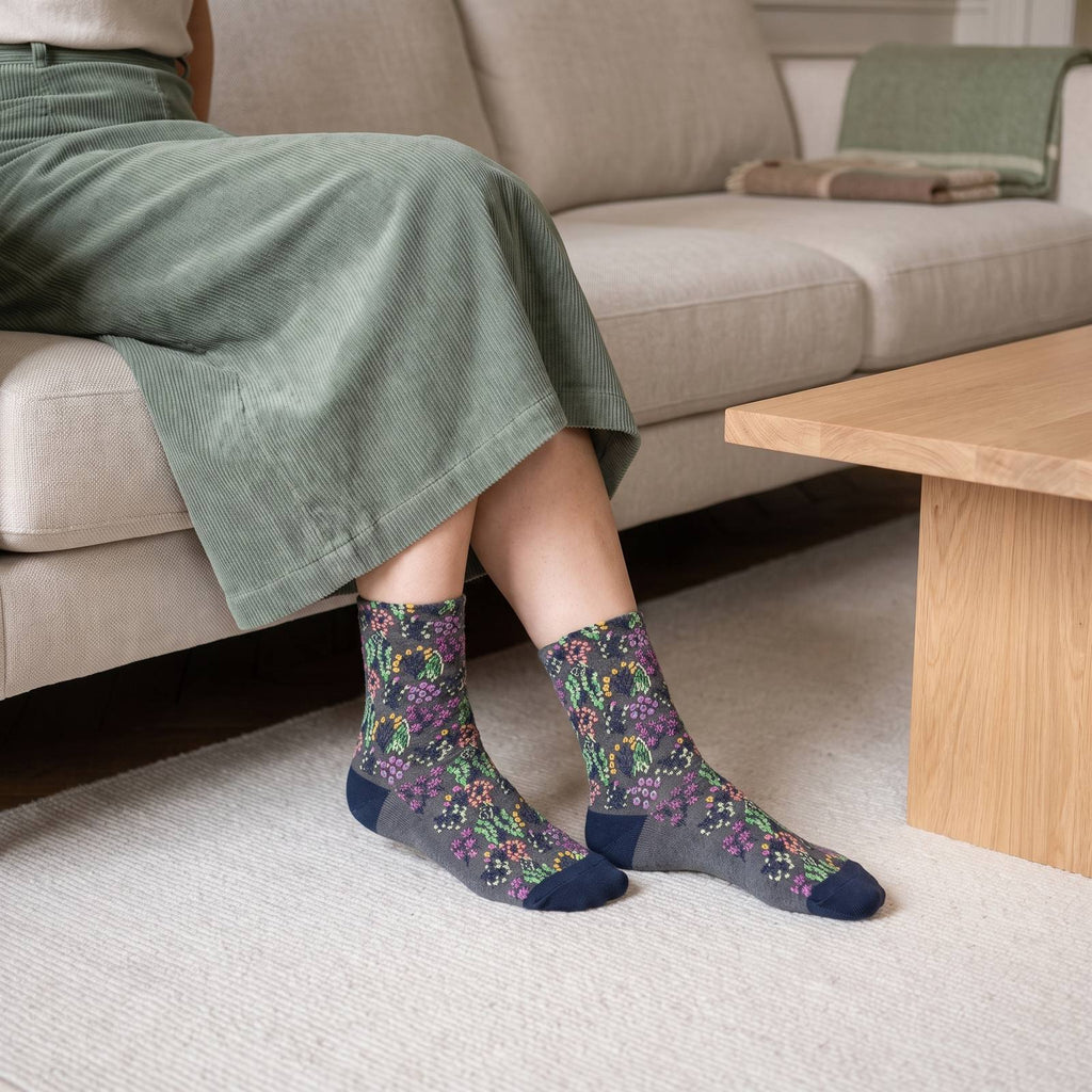 Chaussettes fleuries grises portées, fleurs magenta et vertes et talon bleu marine, pieds posés sur le tapis crème devant un canapé écru