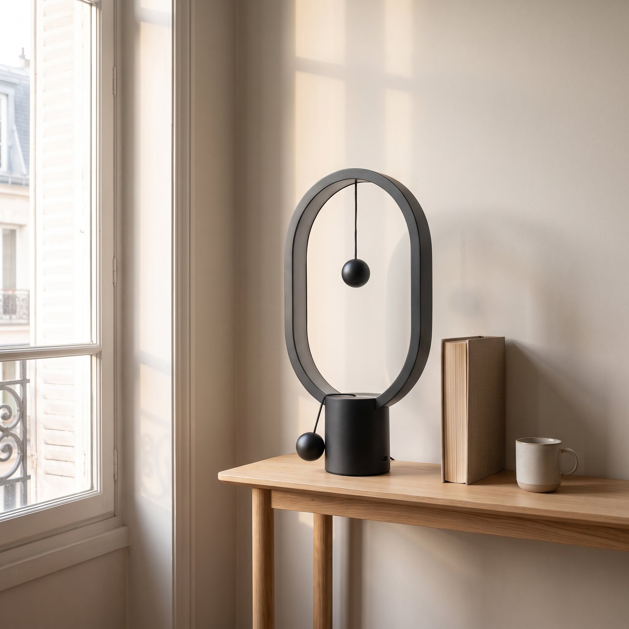 Lampe de chevet ovale en métal noir à billes magnétiques, éteinte en plein jour sur une console, à côté d'un livre Image