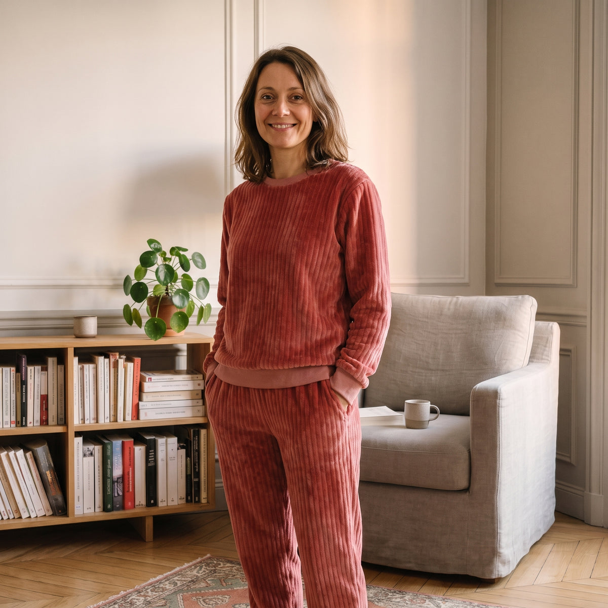Femme debout devant une étagère de livres, une tasse à la main, en pyjama en peluche côtelée rouge brique. Image