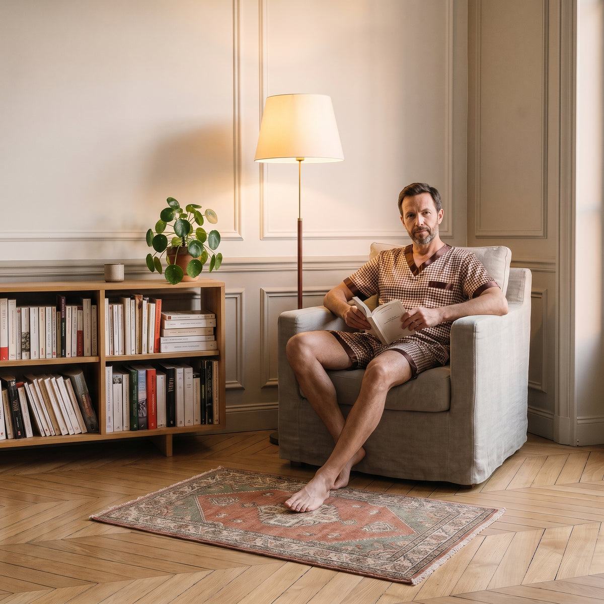 Homme en pyjama de satin pied-de-poule assis dans un fauteuil, un livre ouvert à la main, la lampe allumée Image