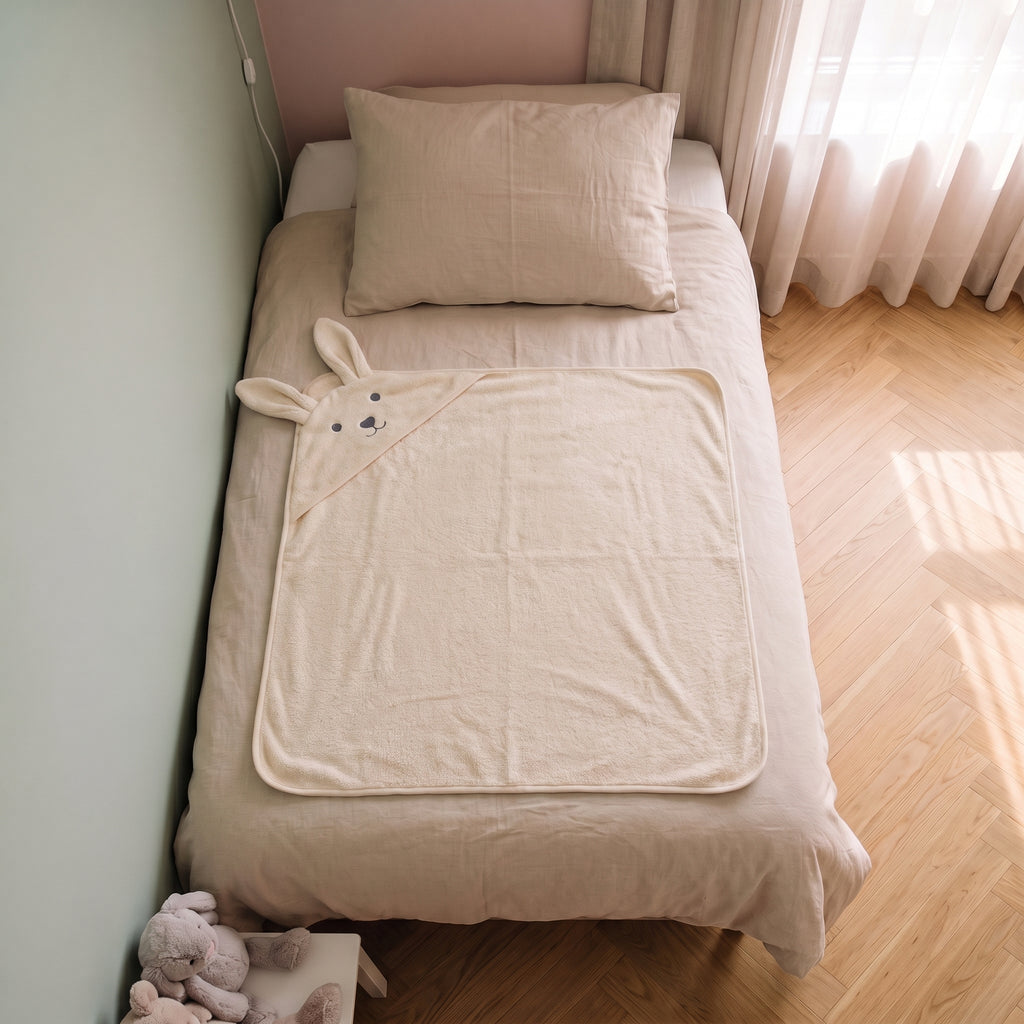 Cape de bain bebe 90 x 90 cm coloris beige, capuche lapin, depliee sur le lit d enfant