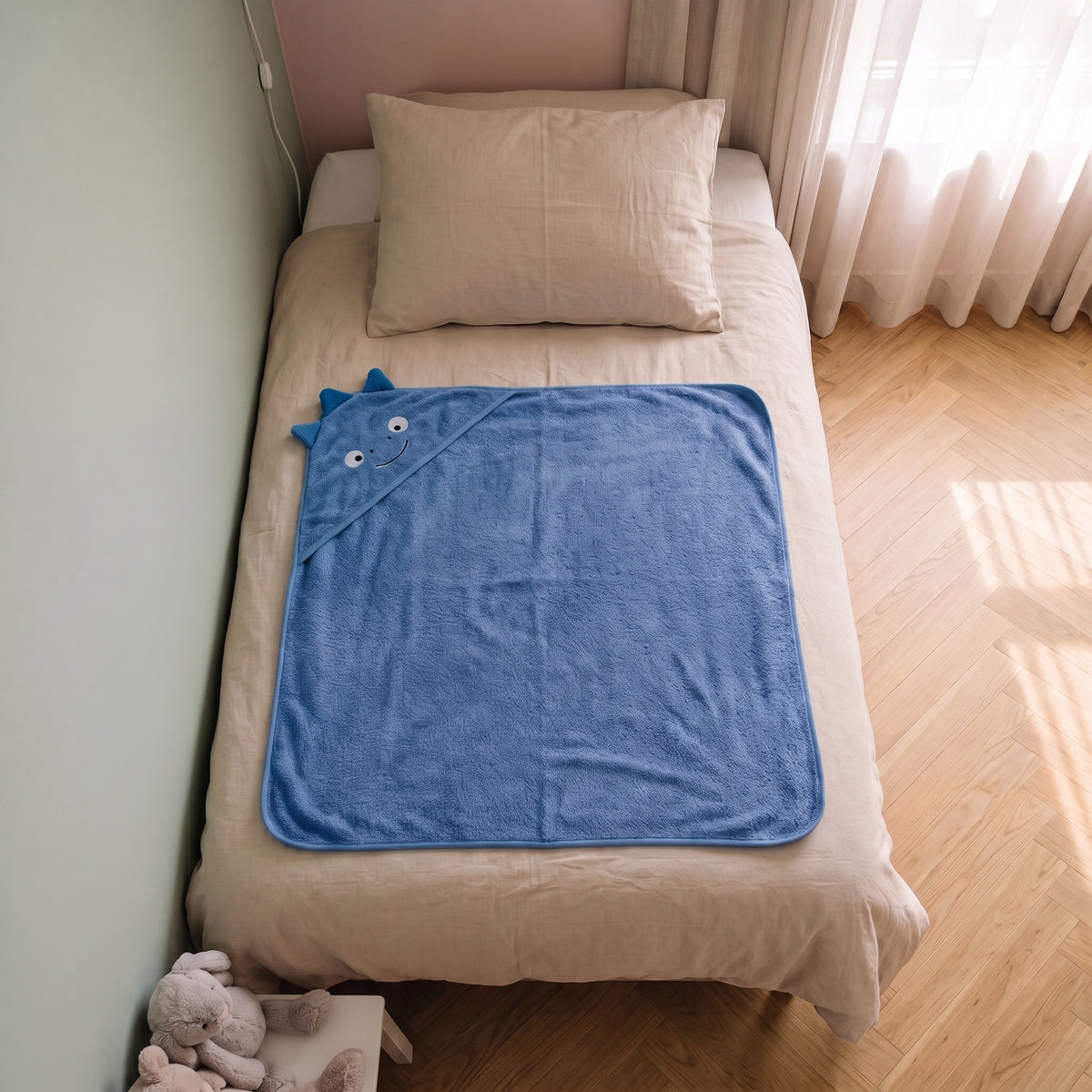 Cape de bain bebe 90 x 90 cm coloris bleu fonce, capuche dinosaure, depliee sur le lit d enfant