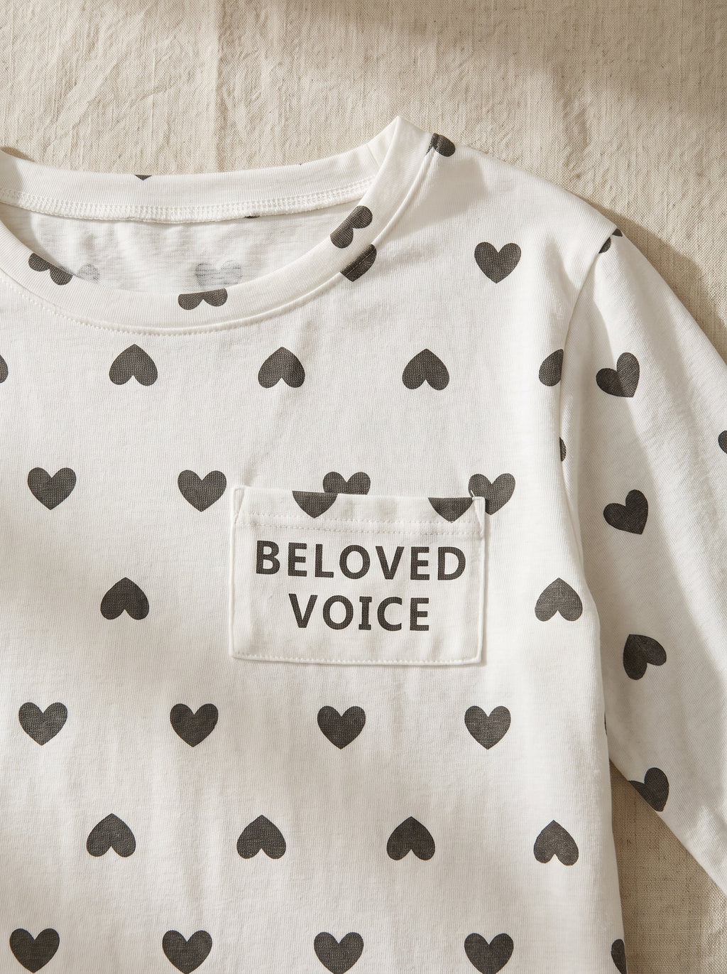 Gros plan du haut blanc seme de petits coeurs gris : la poche poitrine porte les mots Beloved Voice imprimes en gris.