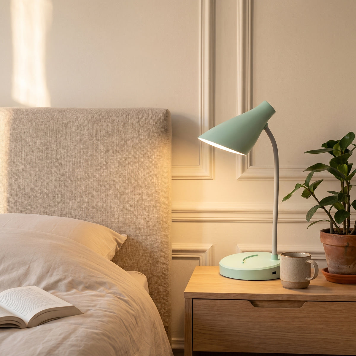 La lampe verte allumée sur une table de chevet en bois clair, à côté d'une tasse en grès, son rond de lumière chaude posé sur le mur de la chambre.