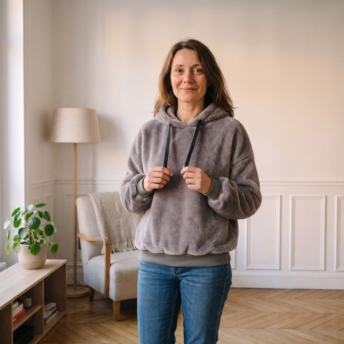 Femme debout dans son coin lecture, en sweat peluche gris taupe et jean, la capuche rabattue dans le dos, les deux brins du cordon noir entre les mains