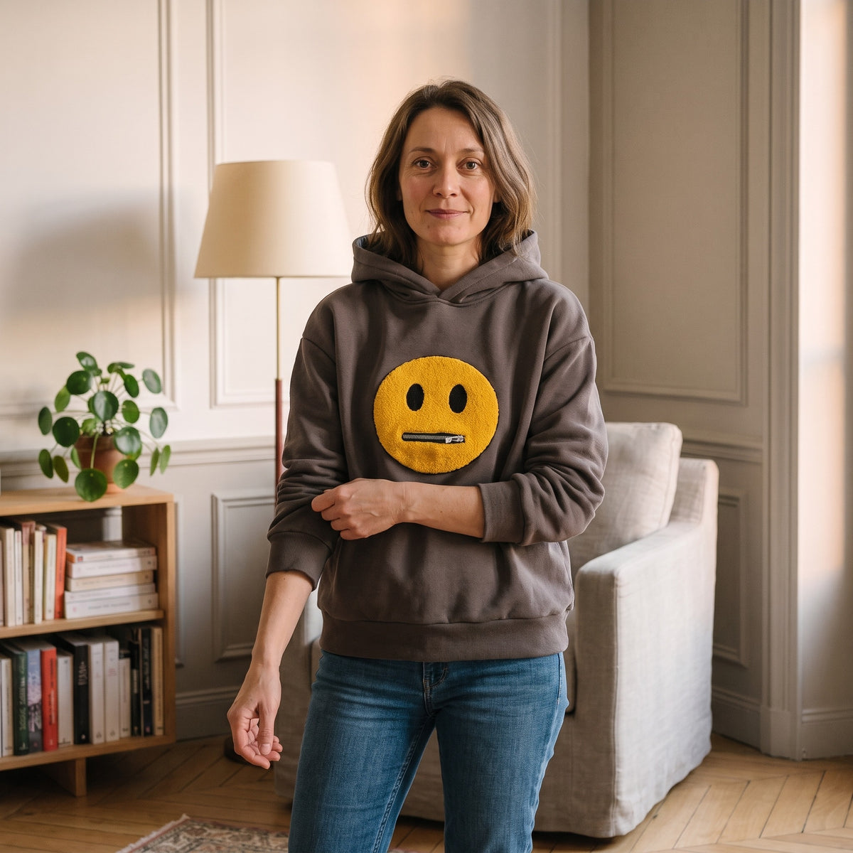 Femme debout dans son salon, en sweat a capuche gris fonce et jean, capuche rabattue dans le dos, le grand smiley jaune en pleine poitrine et la bande cotelee du bas sur les hanches