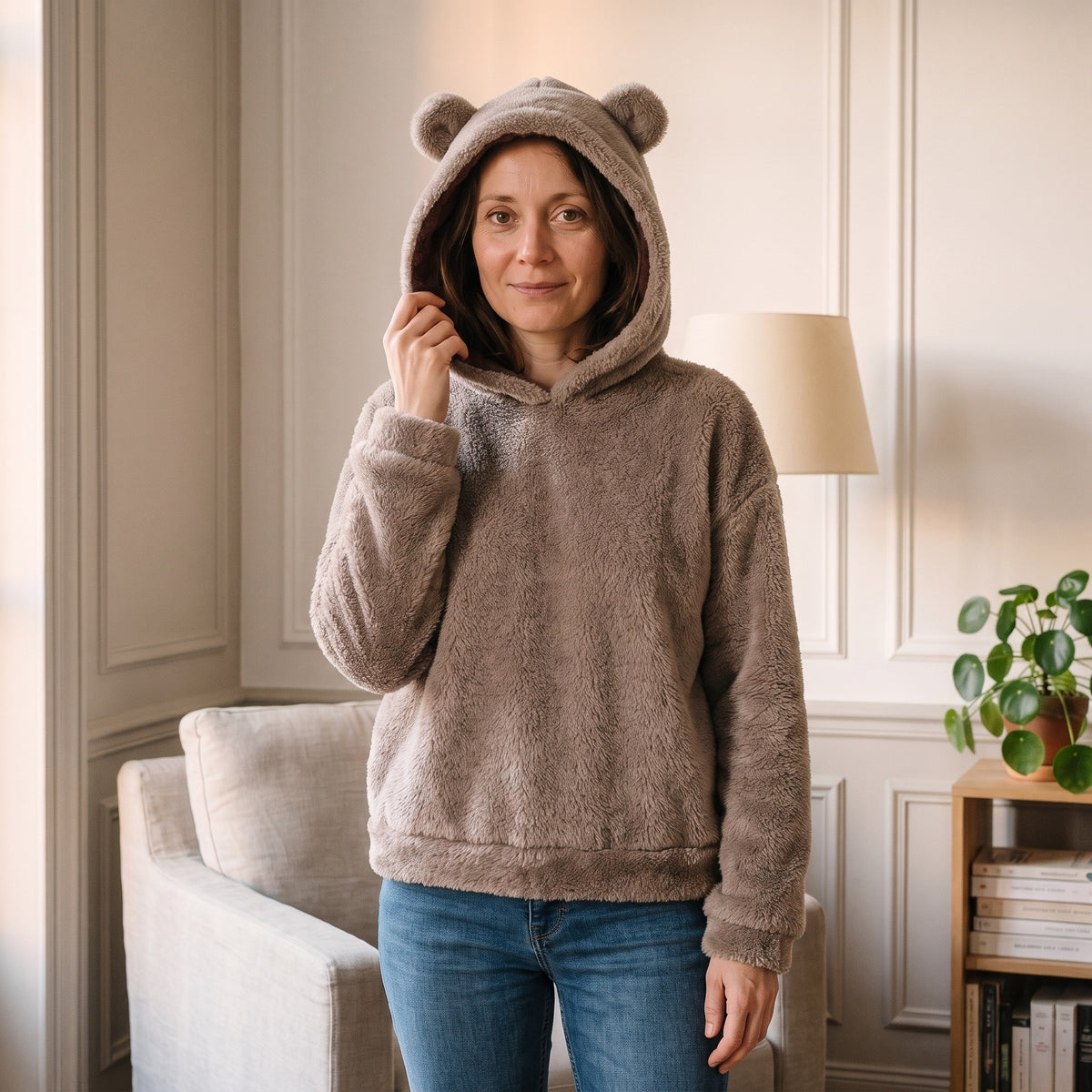 Femme debout dans son coin lecture, en sweat ourson marron taupe et jean, la capuche relevee sur la tete, une main qui en tient le bord pres de la joue