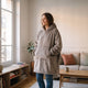 Une femme debout dans son salon haussmannien porte le plaid à capuche gris taupe, les deux mains dans la grande poche du devant, la doublure sherpa crème visible au bord bas