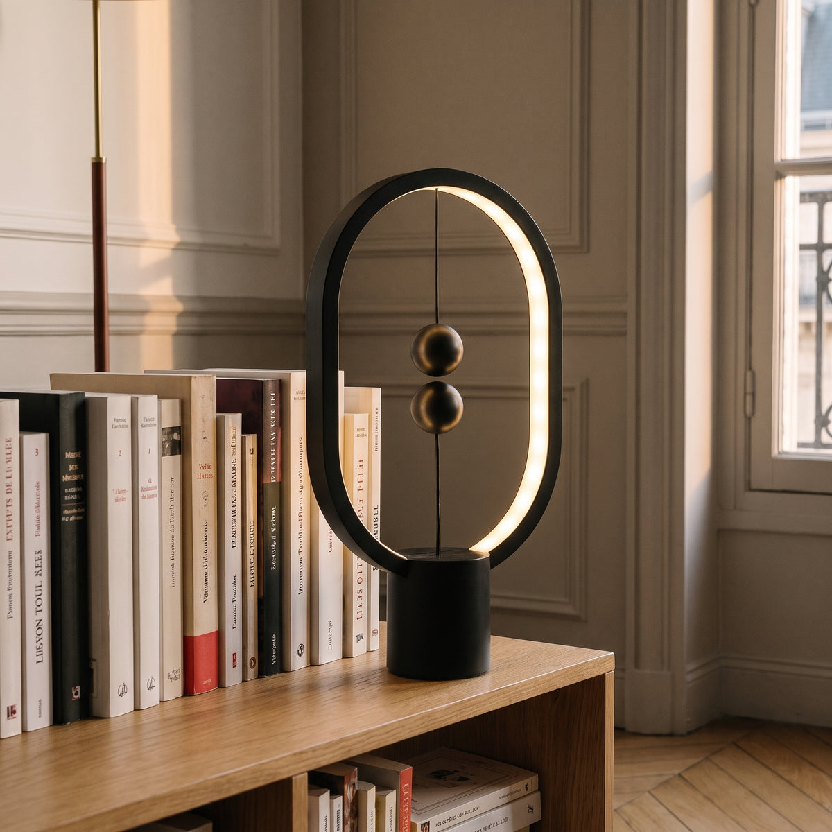 Lampe de chevet ovale en métal noir à billes magnétiques, allumée sur une bibliothèque basse, à côté des livres
