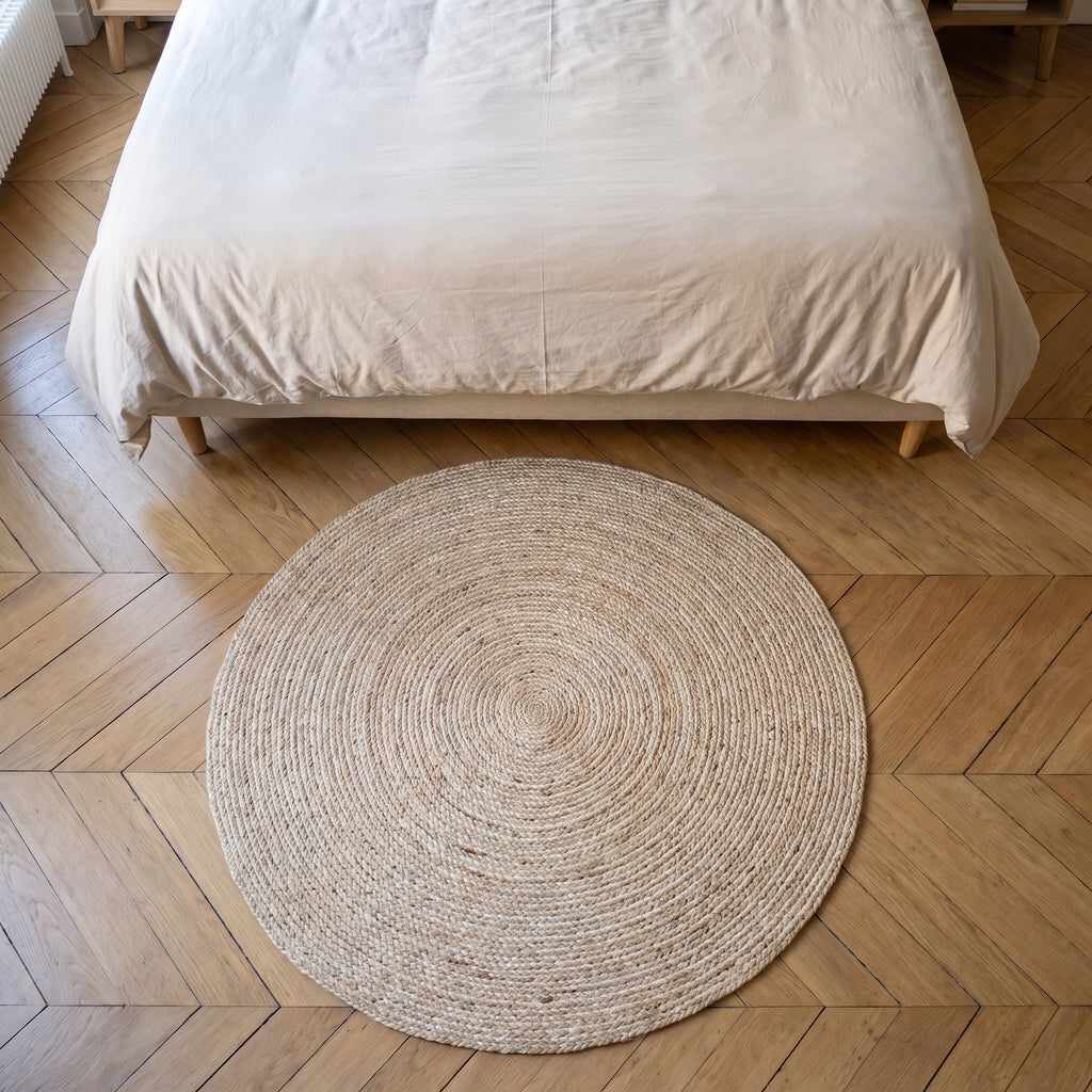 Tapis rond en sisal beige clair vu du dessus, au pied d'un lit, sur un parquet à chevrons