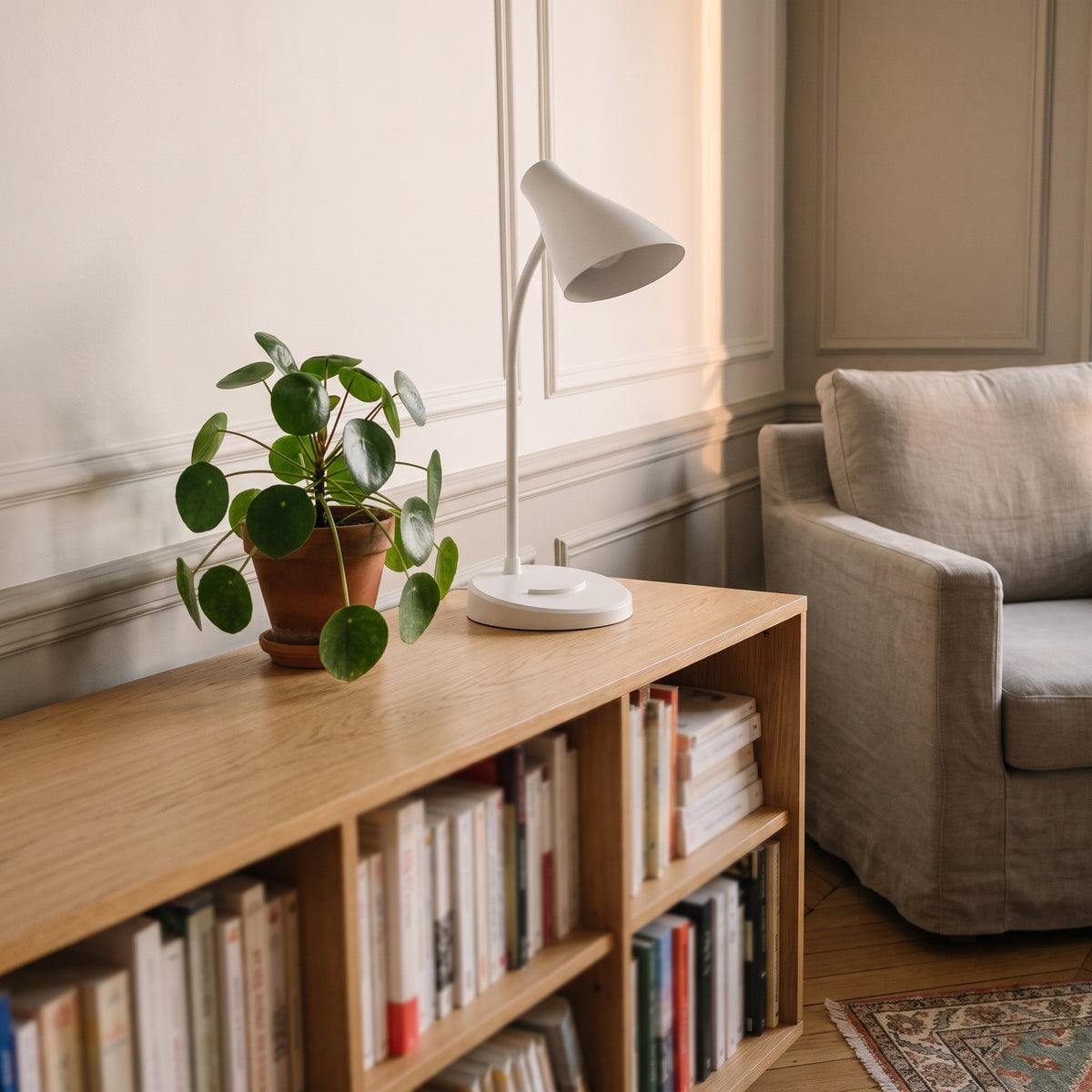 La lampe blanche éteinte sur le plateau d'une bibliothèque basse en chêne, en plein jour, à côté d'un pilea en pot de terre cuite.